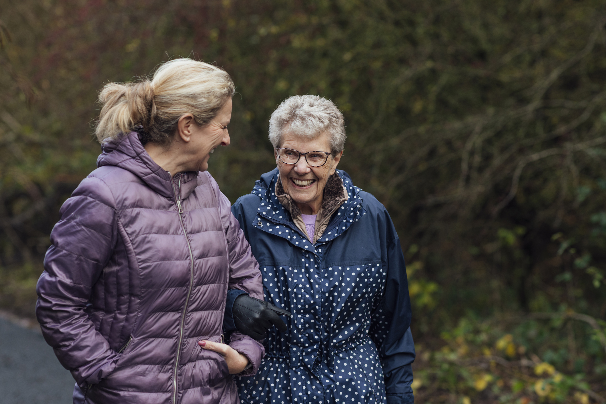 Two Generations on a Stroll A mature woman walking with her mother on a path through a public park while getting exercise in Seghill, Northumberland. The senior woman has dementia and is arm in arm with her daughter for support and care while they look at each other, laugh and enjoy the fresh air.