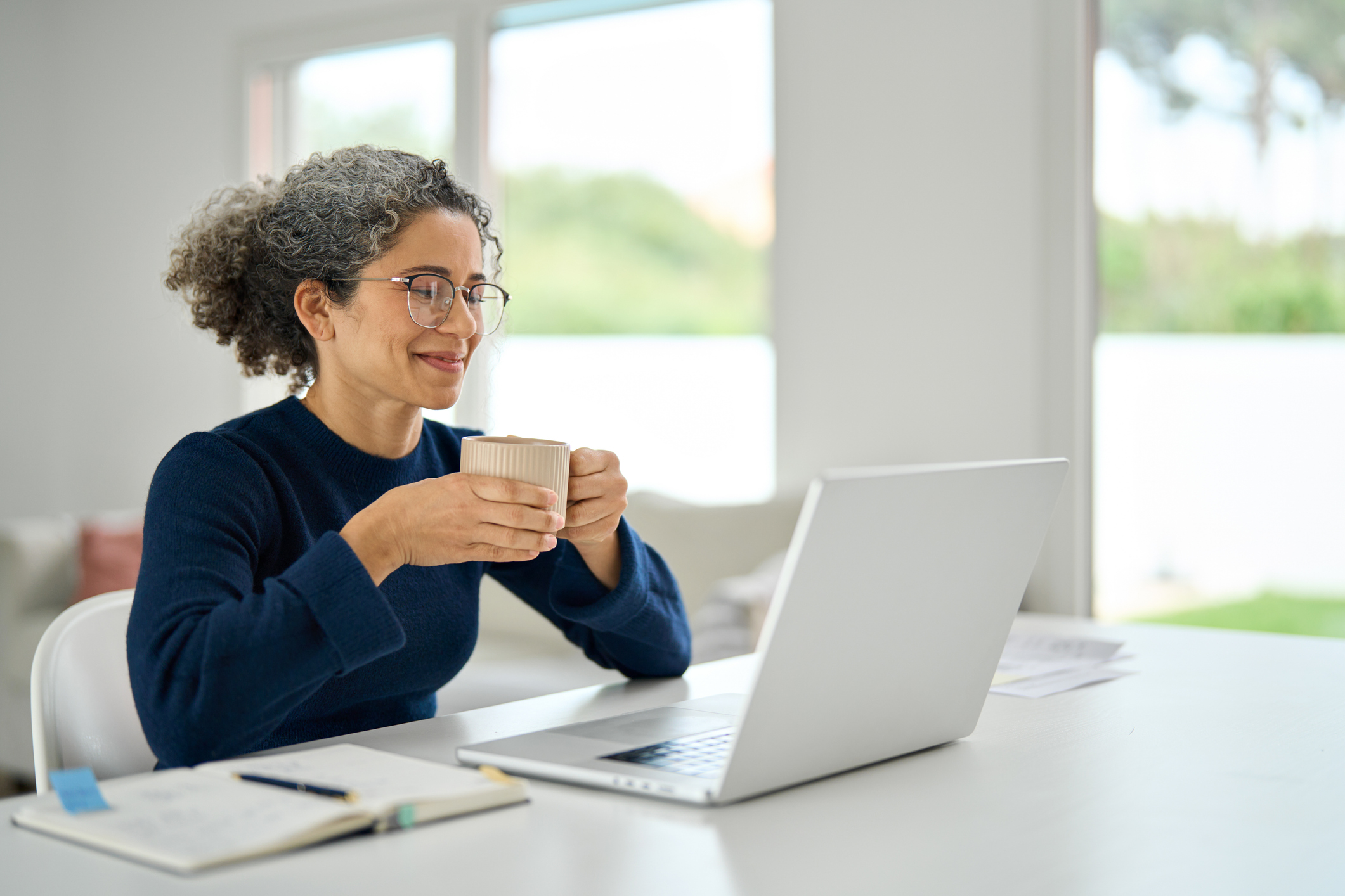 Happy mature woman sitting at home table drinking coffee looking at laptop. Annapolis, MD senior drinking coffee while watching webinar on laptop